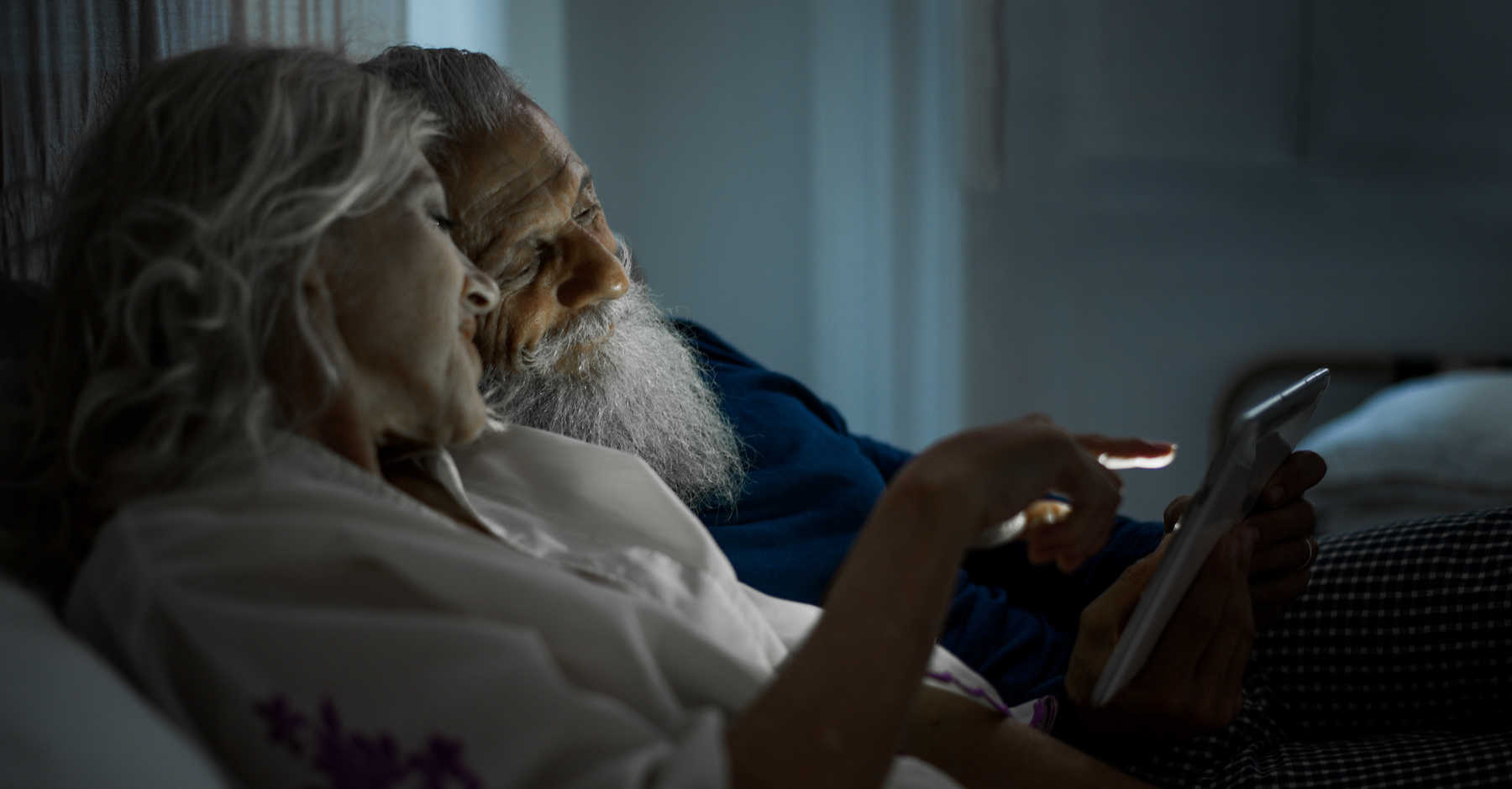 Zufriedenes Seniorenpaar dank Inkontinenzprodukten  kuscheln im Bett und schauen auf ein Tablet
